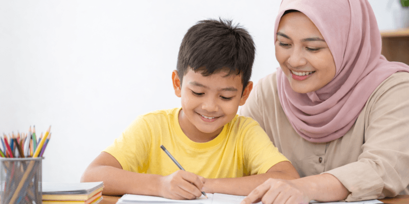 Guru wanita Indonesia berhijab membantu siswa belajar menulis di meja belajar dalam suasana homeschooling yang nyaman di rumah.